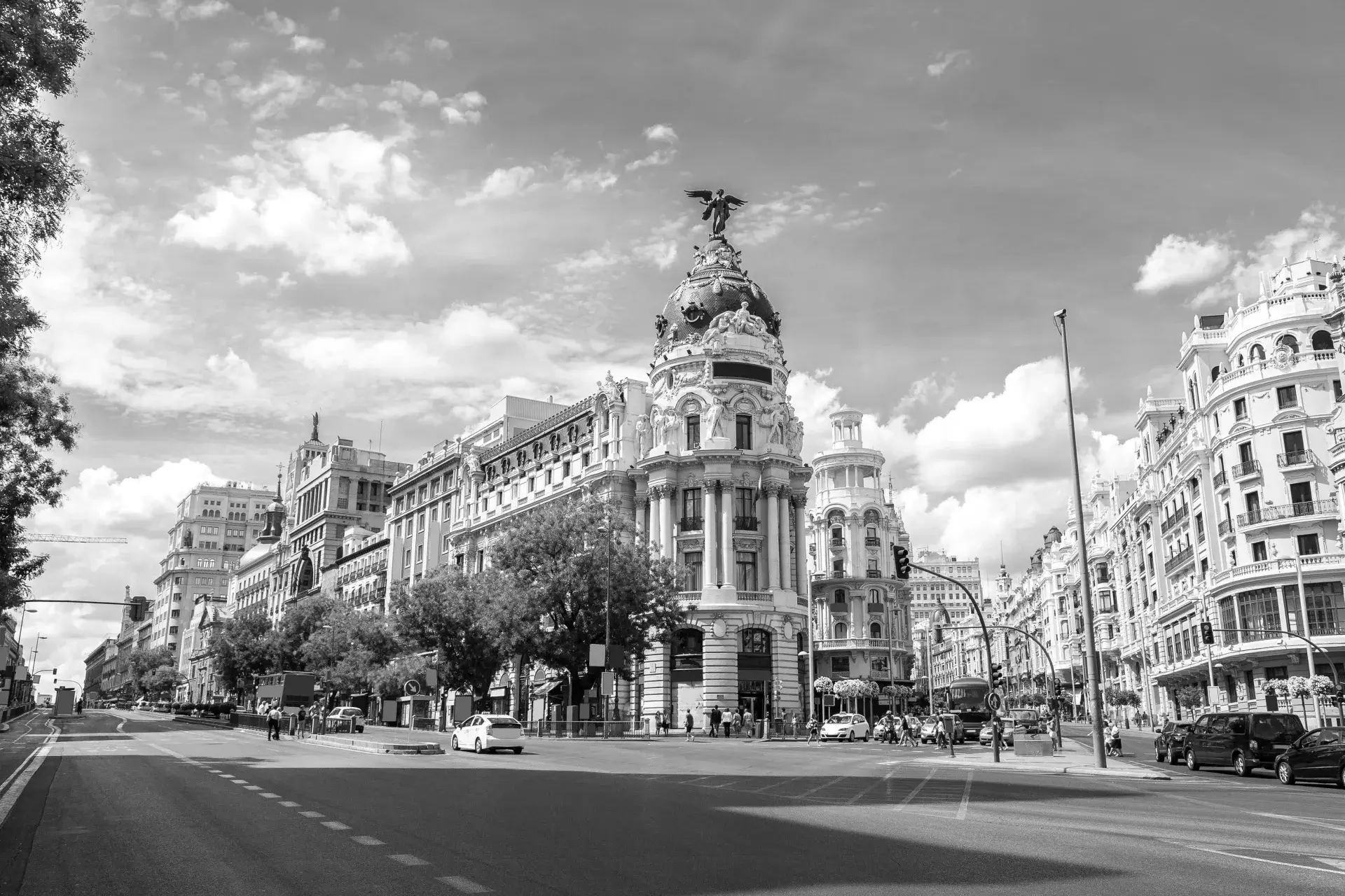 Wide street view of historic buildings with ornate architecture and sculptures under a partly cloudy sky in Madrid.