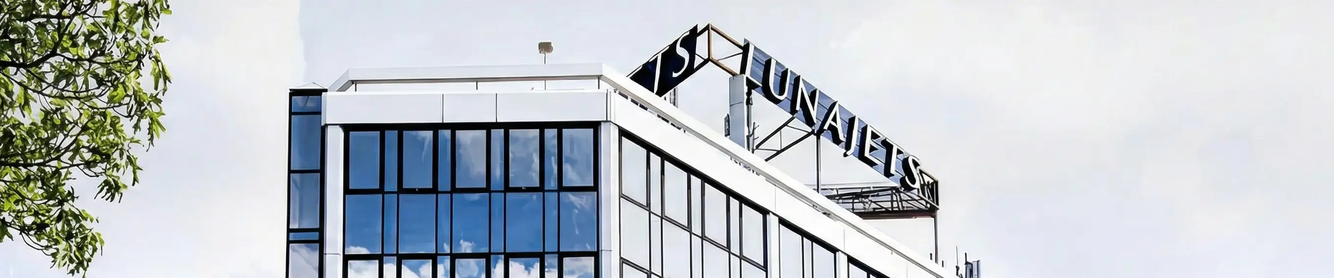 Egy modern irodaház felső sarka fényvisszaverő üvegablakokkal és a tetőn Lunajets táblával, részben faágakkal eltakaró.