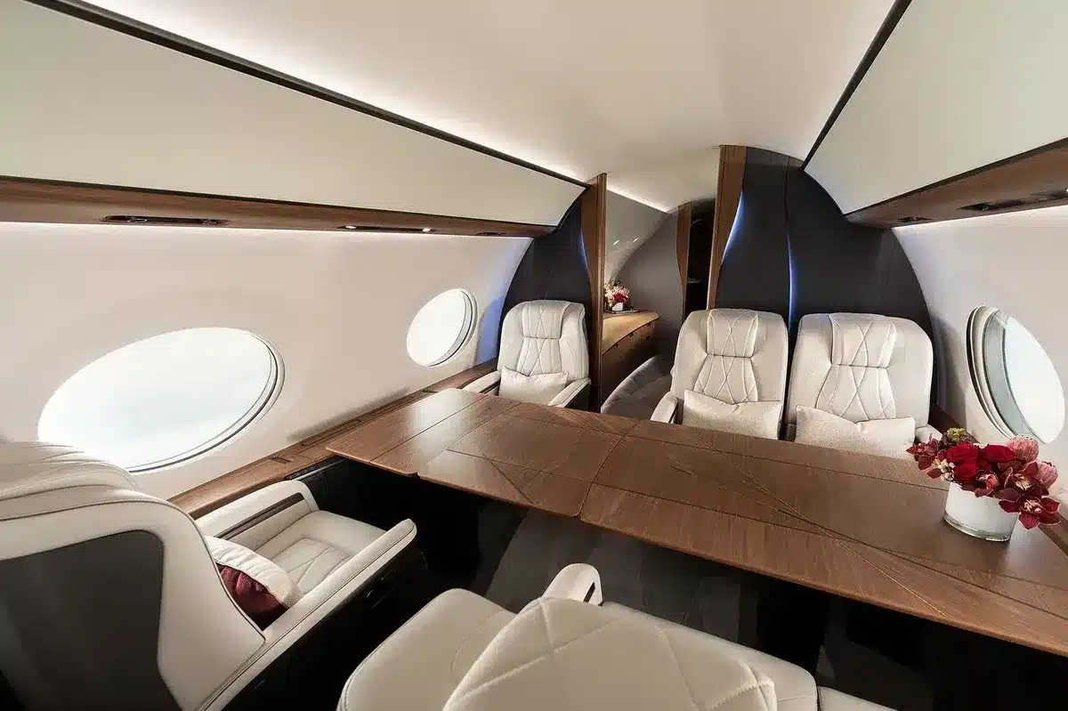 Luxurious private jet cabin with cream leather seats, a wooden table, large oval windows, and a vase of red flowers.