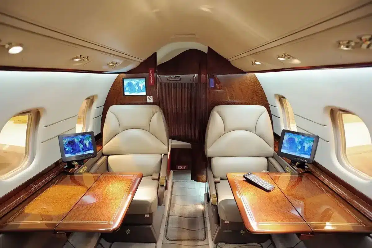 Interior of a private jet cabin with two beige leather seats facing fold-out wooden tables, small display screens, and windows on both sides.