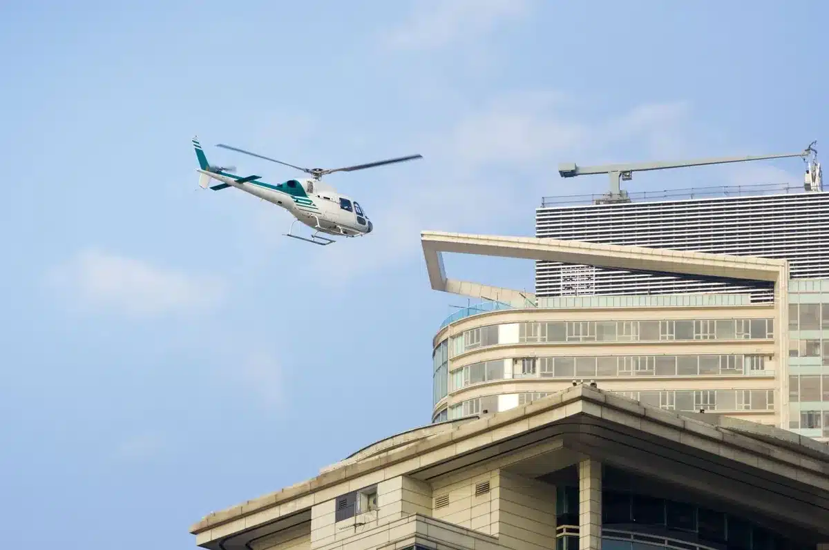 White and teal helicopter flying near a modern building under a blue sky.