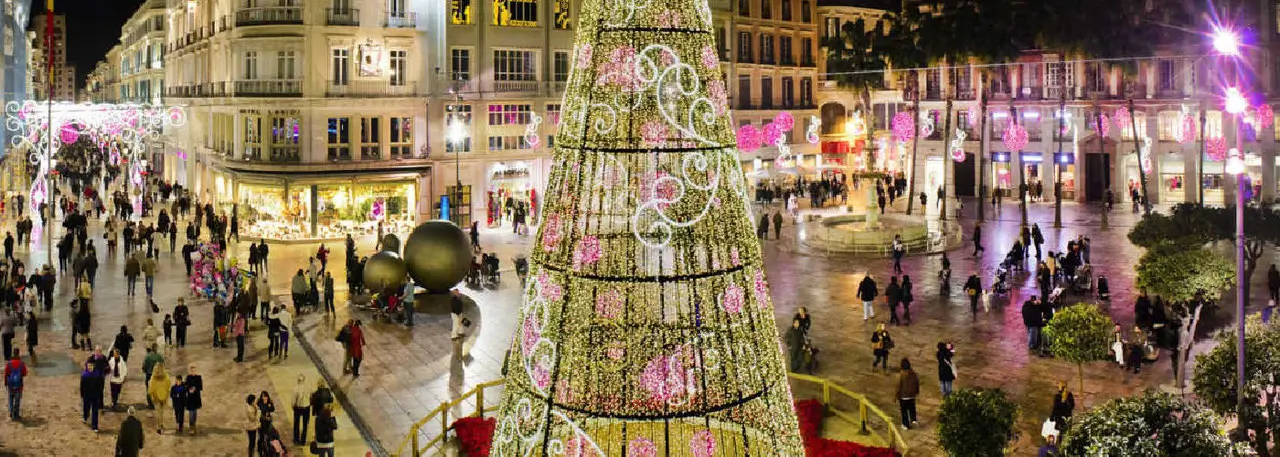Nighttime view of a busy plaza decorated with a large illuminated Christmas tree and festive pink and white lights, surrounded by people and historic buildings.