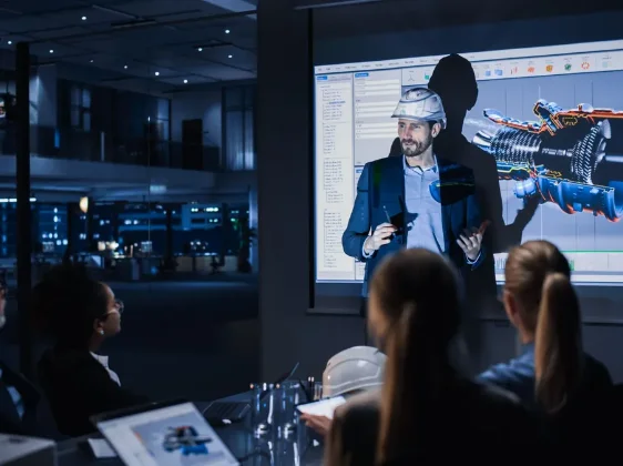 Engineer wearing a hard hat giving a presentation on a mechanical device to colleagues in a modern office.