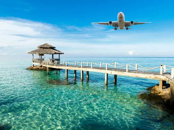 Airplane flying over a wooden pier with a thatched gazebo above clear turquoise ocean water.