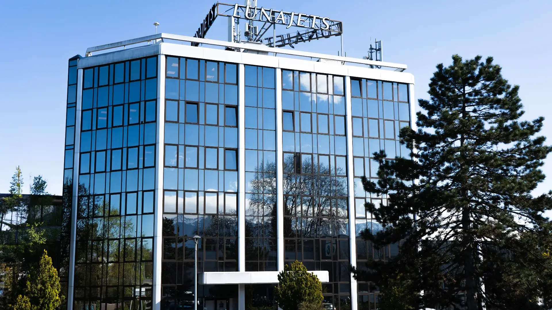 Modern üveg irodaház nagy ablakokkal tükröződő fákkal és eggel, valamint Lunajets táblával a tetőn.