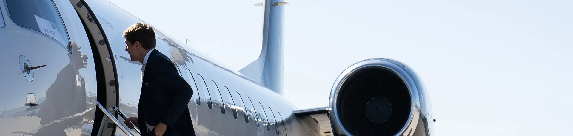 Uomo in giacca e cravatta a bordo di un jet privato con motore e coda visibili in una giornata limpida.