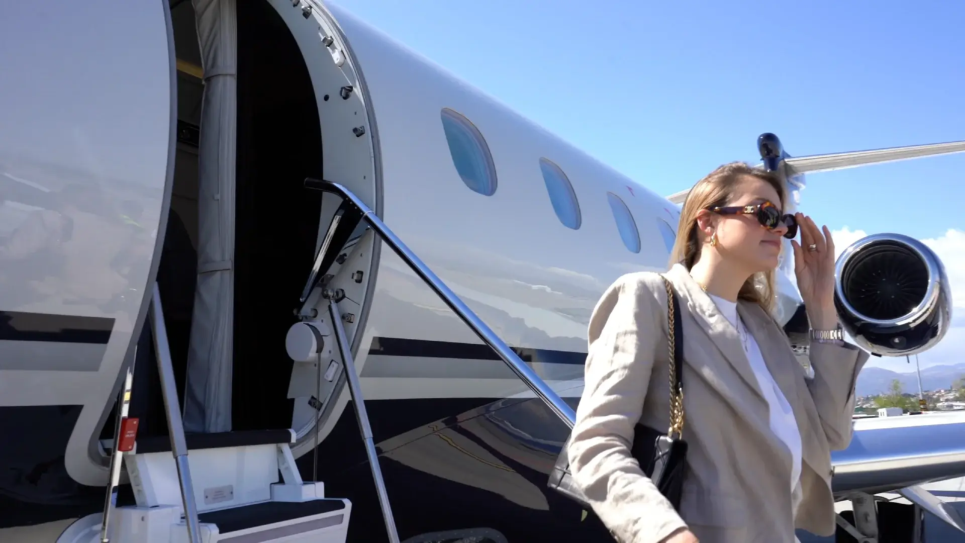 A woman wearing sunglasses and a beige blazer walking down the stairs of a private jet on a sunny day.