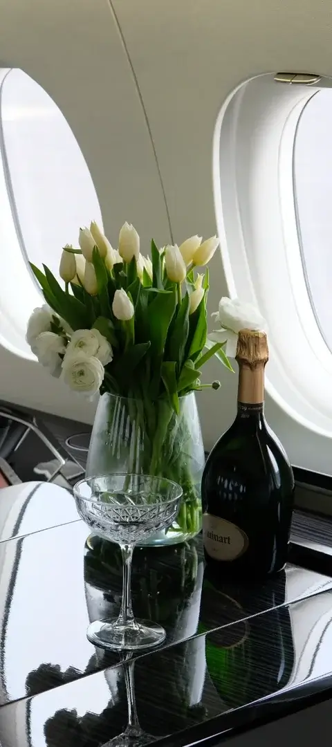 Private jet interior with leather seats, a black glossy table holding two crystal champagne glasses, a vase of white tulips, and a bottle of champagne.