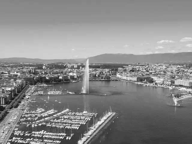 Aerial view of a Geneva lake with numerous docked sailboats, a prominent water fountain jet in the center, and a cityscape with mountains in the background.