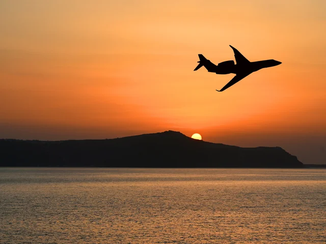 Silhouette eines Privatjets, der bei Sonnenuntergang über das Meer fliegt, wobei die Sonne hinter einem fernen Hügel teilweise sichtbar ist.
