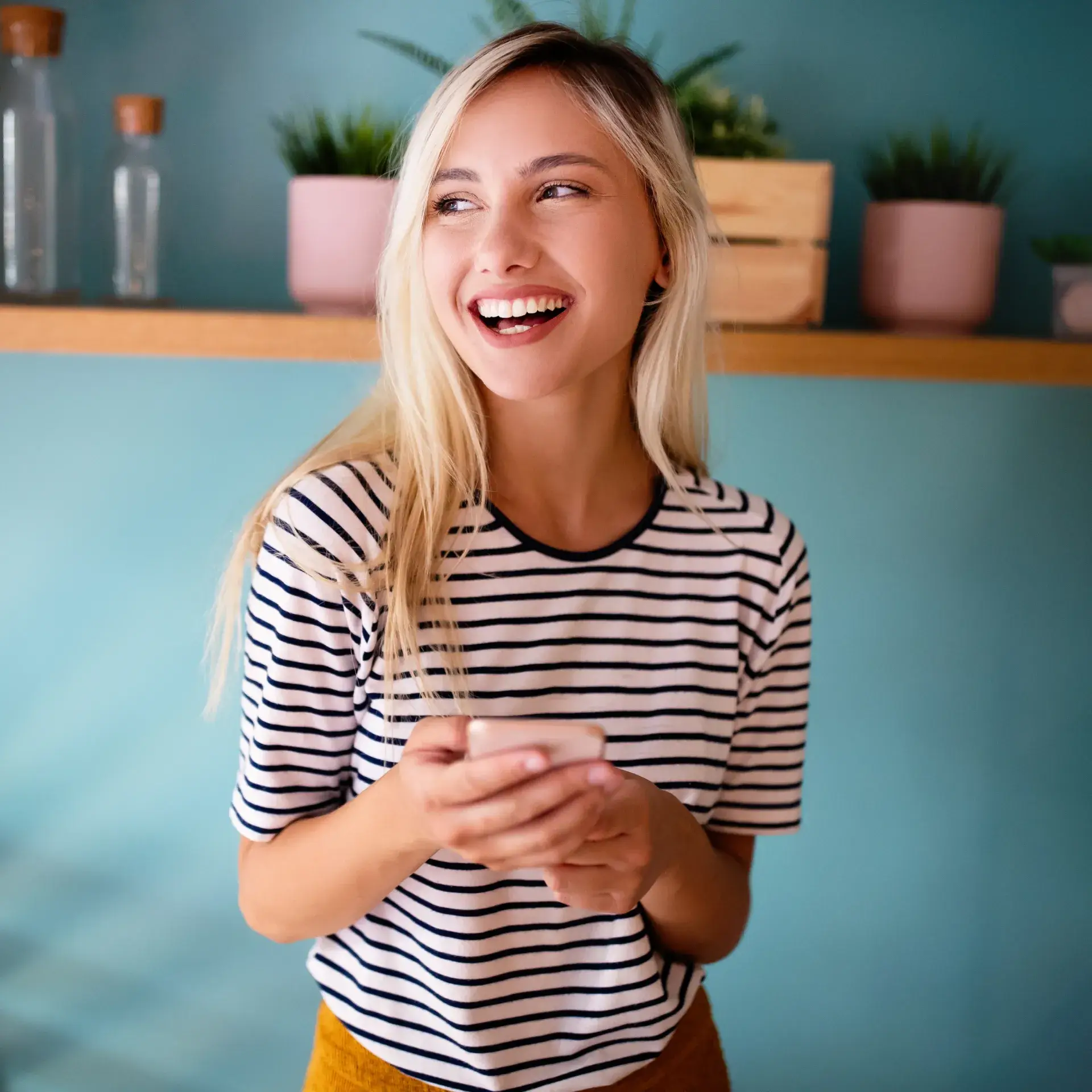 Photo of a smiling woman with long blonde hair, wearing a striped shirt, holding a phone, in front of a blue wall with plants.