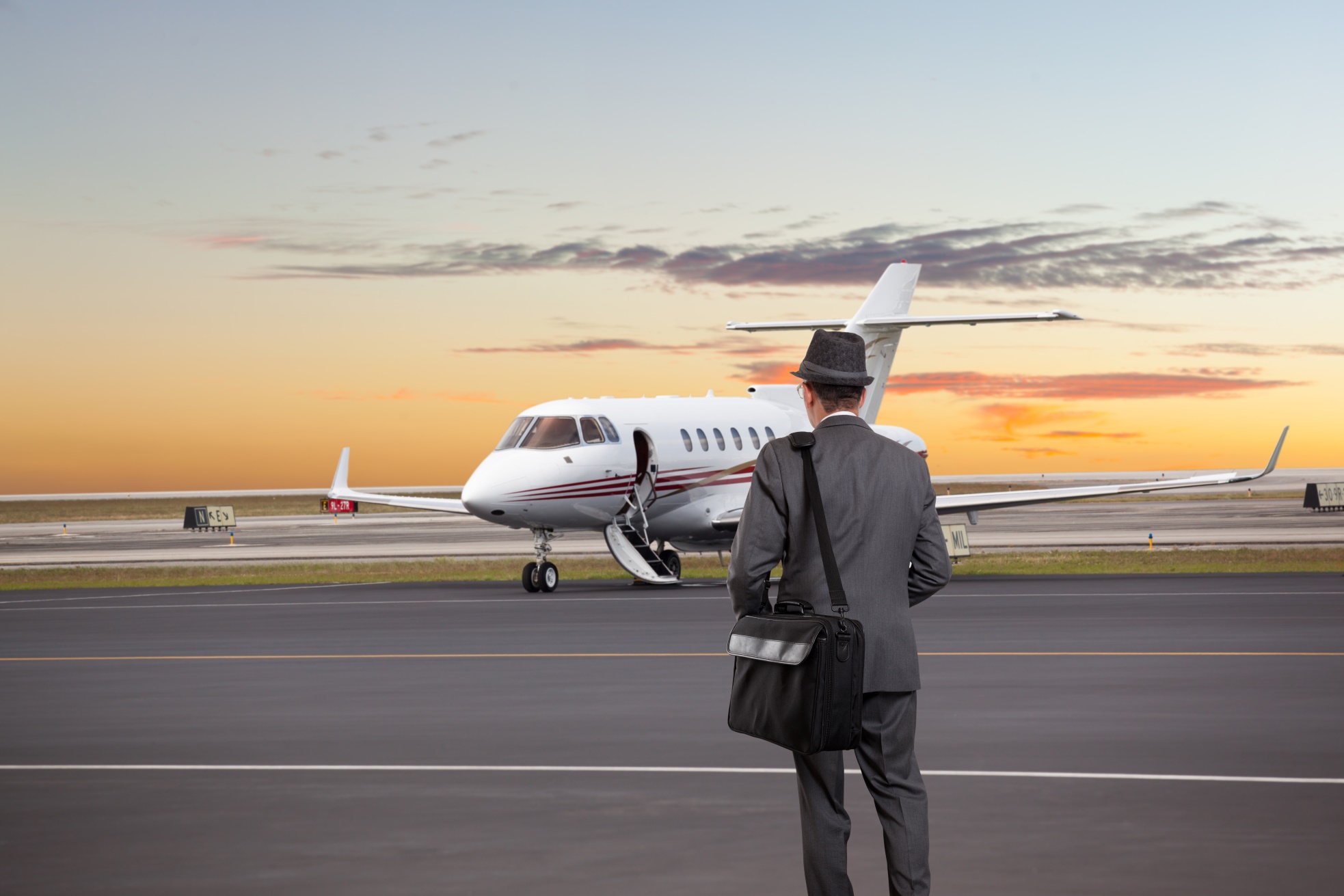 Mężczyzna w szarym garniturze i kapeluszu zmierzający w stronę prywatnego odrzutowca o zachodzie słońca