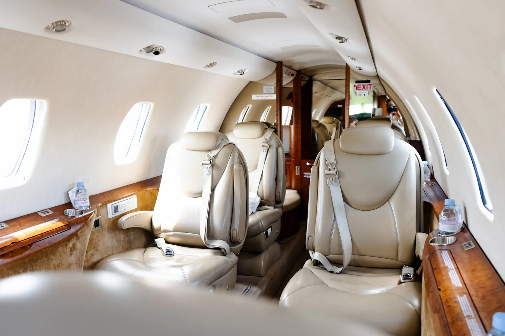 Interior cabin view of Cessna Citation XLS with cream leather seats, wooden trim, windows, and exit sign visible.