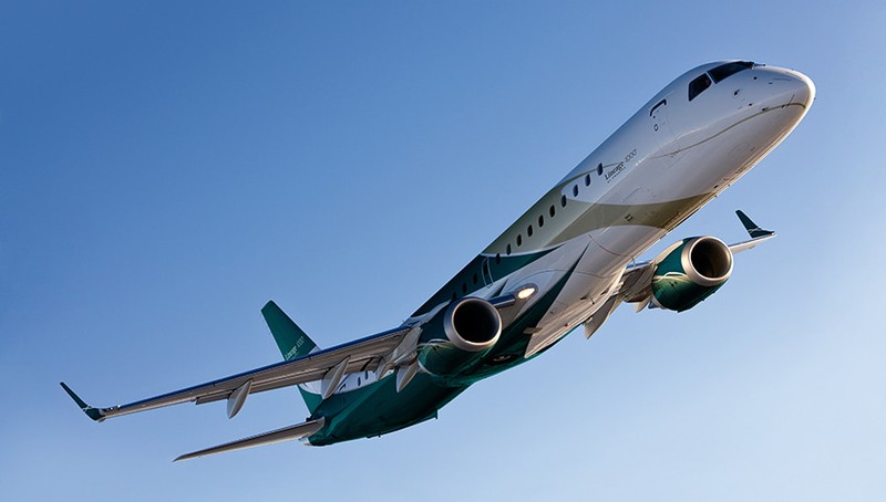 Embraer Lineage 1000 private jet in flight against clear blue sky, angled upward view showing twin engines and teal tail fin.