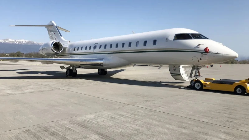 Bombardier Global 6000 luxury jet parked on tarmac with ground equipment, mountains visible in background, side profile view.