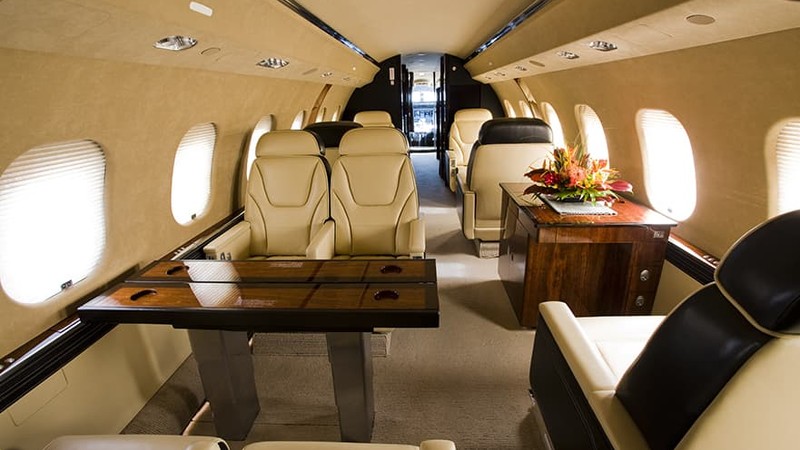 Bombardier Global 6000 cabin interior with cream leather seats, wooden tables, large windows, and beige walls.