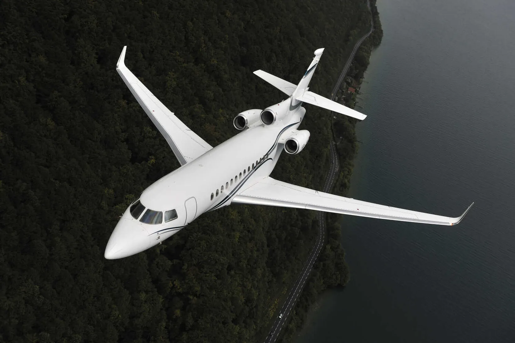 White Dassault Falcon 7X private jet in flight over forested landscape with road below, aerial overhead view.