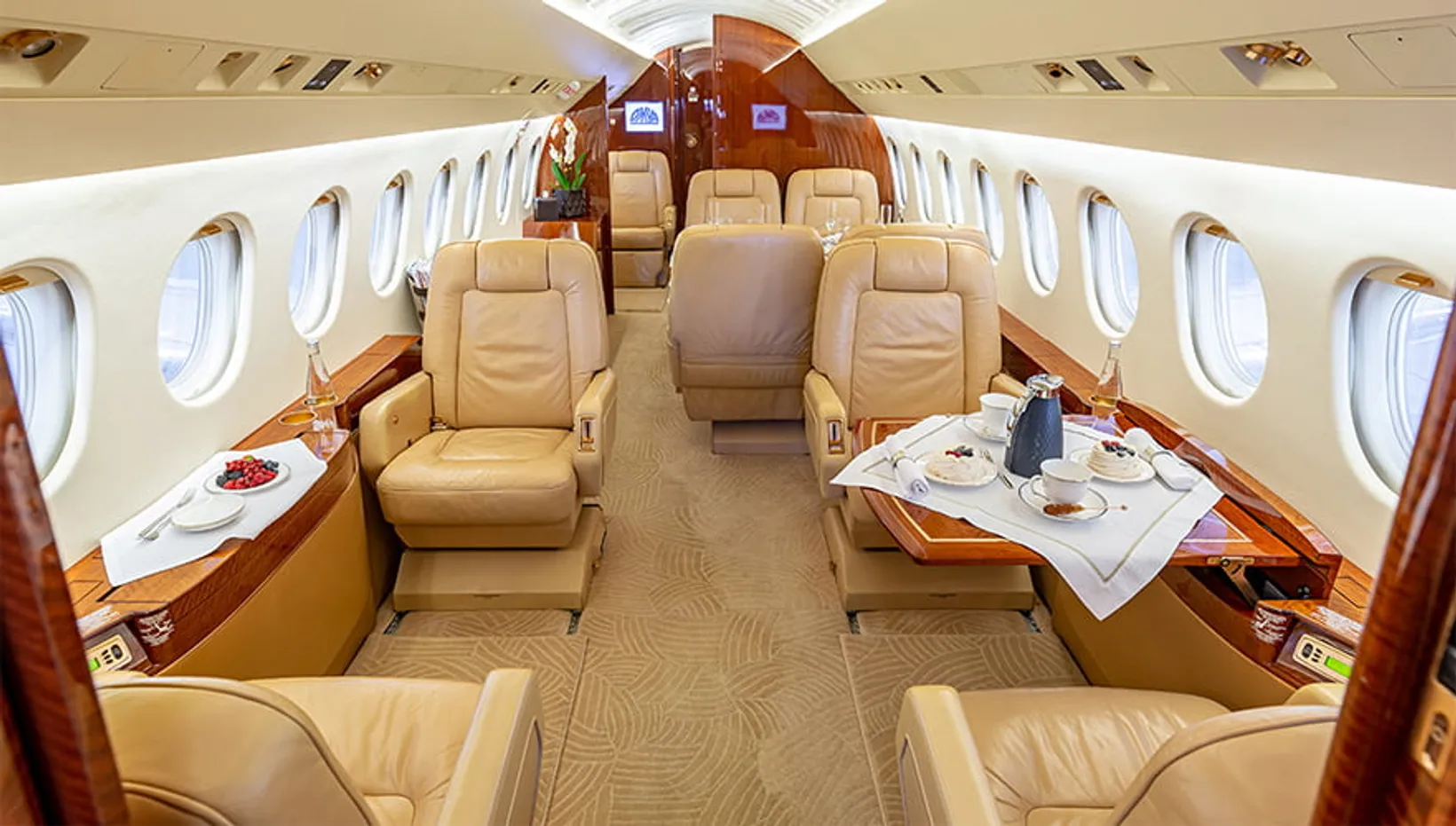 Interior cabin view of a Dassault Falcon 900C private jet showing tan leather seats, wooden trim, dining tables with place...
