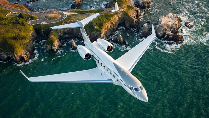 Gulfstream G600 private jet in flight over coastal cliffs and turquoise water, aerial top-down view showing twin engines a...