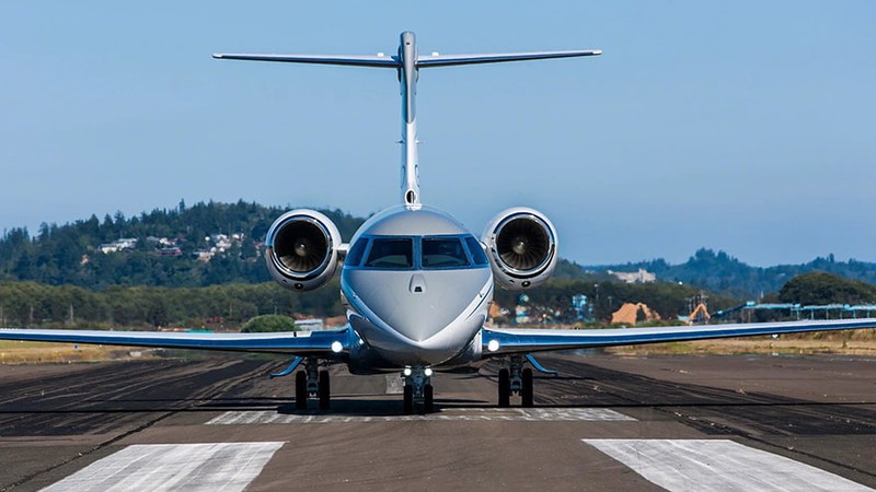 Gulfstream G280 twin-engine jet parked on runway, front view, with forested hills and clear sky in background.