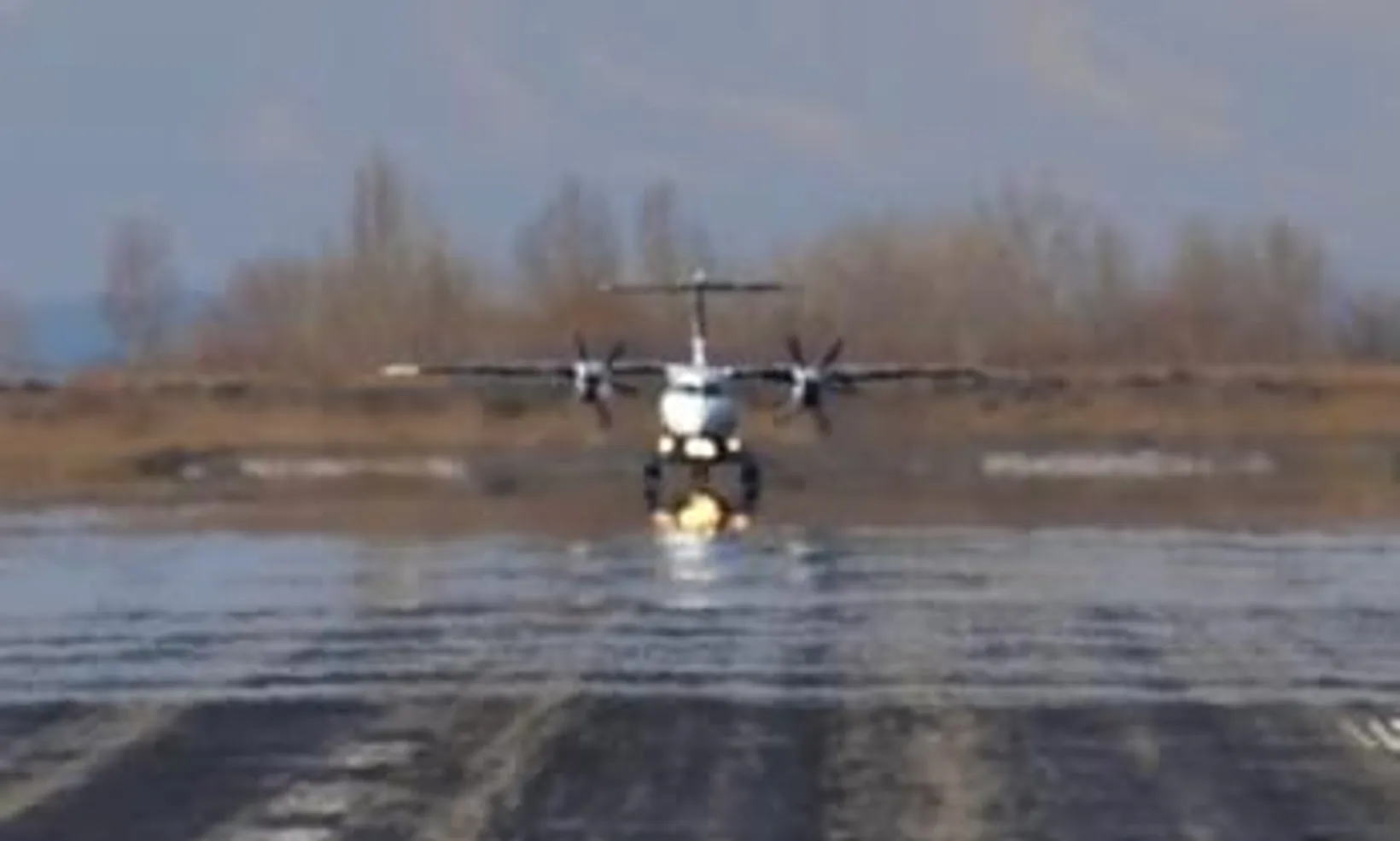 Dornier 328 twin-engine aircraft landing on wet runway with headlights on, misty forest backdrop, front-facing view.