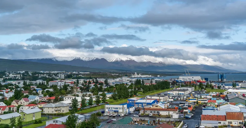 Panorámás kilátás nyílik az izlandi Akureyrire, bemutatva a város színes épületeit, ipari kikötőjét és a környező hófödte hegyeket.