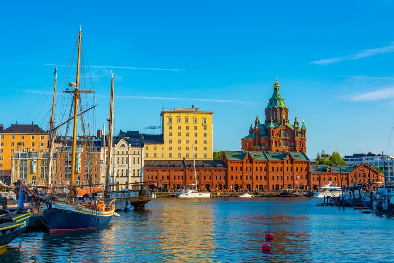 Tętniący życiem widok na nabrzeże Helsinek, prezentujący zabytkowe budynki miasta, łodzie i kultową Katedrę Uspieńską pod czystym, błękitnym niebem.