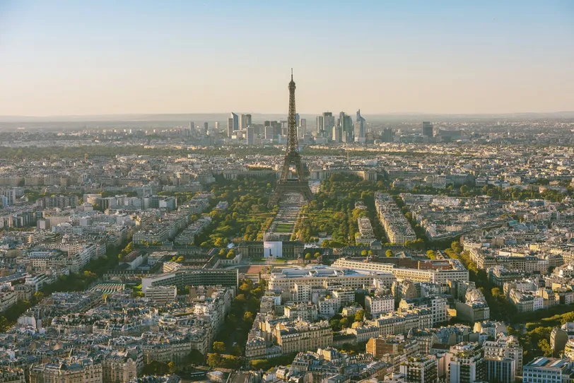 Vista aerea mozzafiato di Parigi, con l'iconica Torre Eiffel in mezzo all'ampio paesaggio urbano, con il moderno skyline di La Défense sullo sfondo, sotto un cielo azzurro e limpido.
