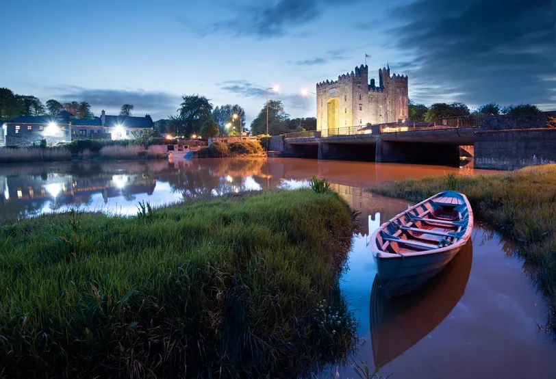 Varázslatos éjszakai kilátás a Bunratty kastélyra, tükröződik a nyugodt folyóvízben, előtérben egy hagyományos csónakkal.