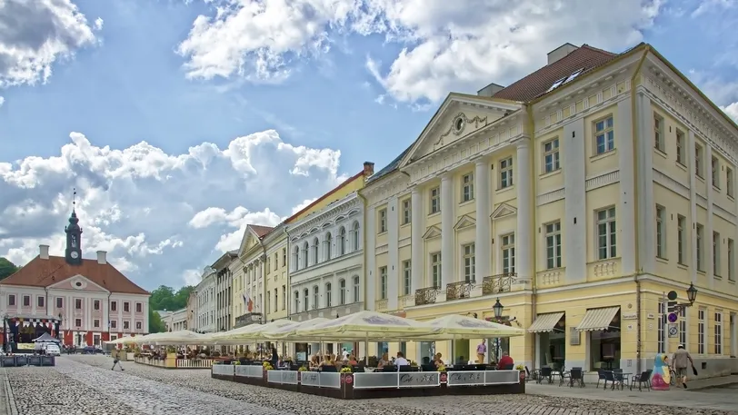 Vivace piazza del municipio di Tartu, in Estonia, con caffè all'aperto, edifici storici e l'iconico municipio di Tartu sotto un cielo parzialmente nuvoloso.