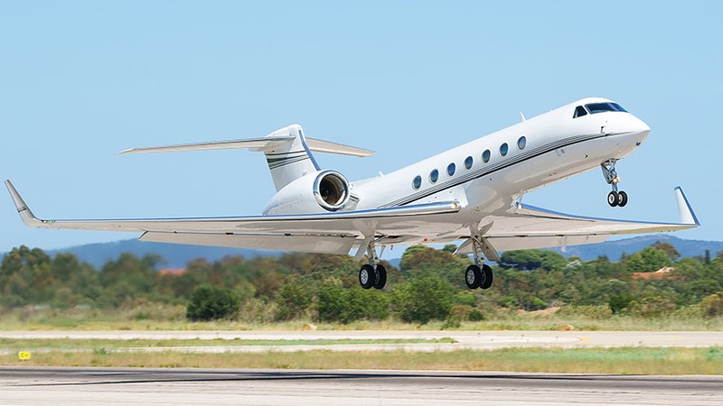 White Gulfstream GV private jet taking off from runway with landing gear down, mountains visible in background.