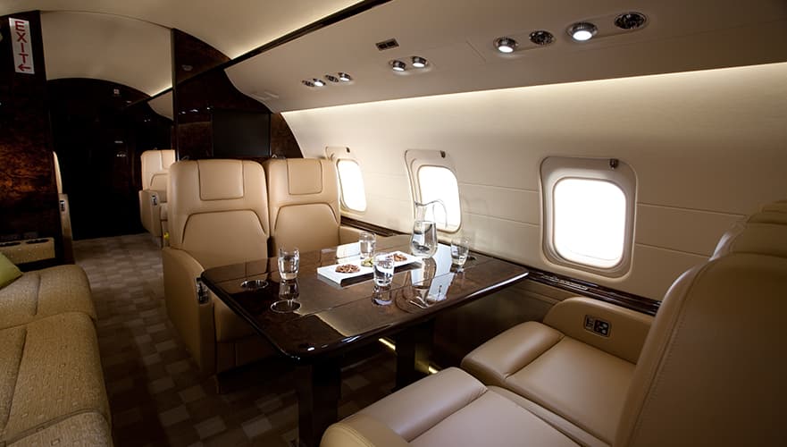 Bombardier Challenger 850 cabin interior with cream leather seating, black conference table, windows, and overhead lighting.
