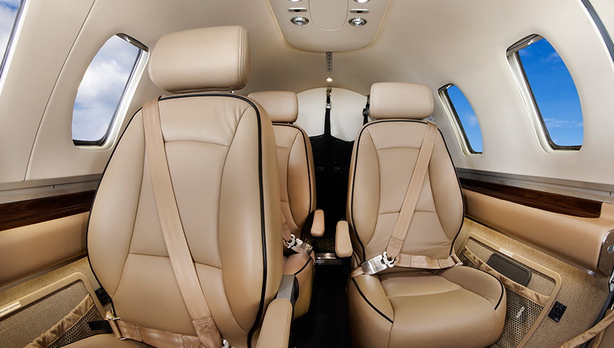 Interior cabin view of Eclipse 550 private jet showing tan leather seats, windows with blue sky, overhead lighting, and wo...