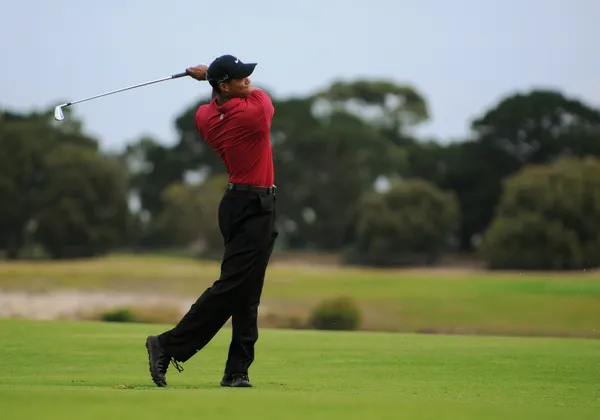 Golfista w czerwonej koszulce kończący zamach na zielonym fairwayu z drzewami w tle