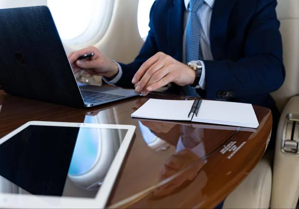 Pasażer pisze na laptopie w prywatnym odrzutowcu, na stole stoi kawa i notatnik