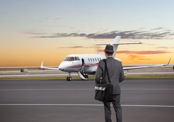 Mężczyzna w szarym garniturze i kapeluszu zmierzający w stronę prywatnego odrzutowca o zachodzie słońca