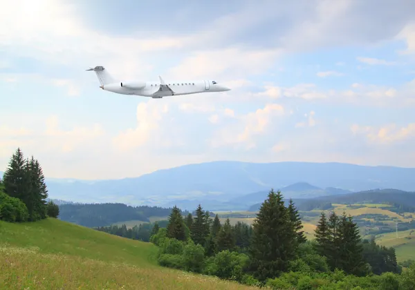 Biały odrzutowiec biznesowy lecący nad zielonymi górami i lasami pod błękitnym, zachmurzonym niebem