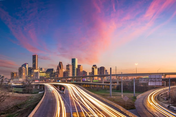 Skyline de Houston au coucher de soleil avec des traînées lumineuses sur les autoroutes courbes et les gratte-ciel du centre-ville reflétés dans la lumière de l'heure d'or