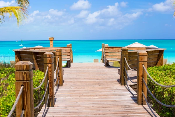 Plage turquoise des Caraïbes avec une promenade en bois, des parasols blancs et du sable pristine menant à des eaux cristallines