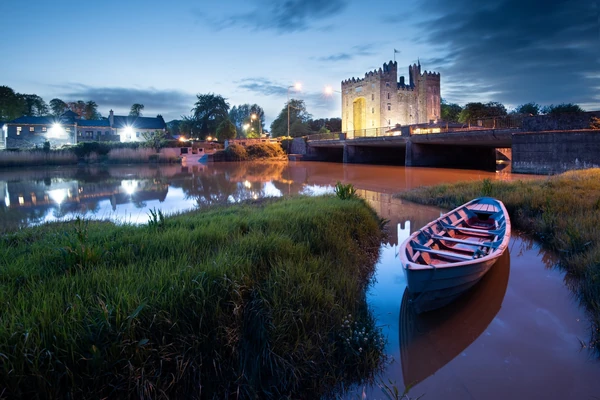 Una encantadora vista nocturna del castillo de Bunratty, que se refleja en las tranquilas aguas del río con un barco tradicional en primer plano.