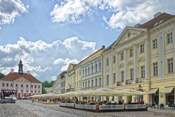 Place de la mairie vibrante à Tartu, en Estonie, avec des cafés en plein air, des bâtiments historiques et l'emblématique mairie de Tartu sous un ciel partiellement nuageux.