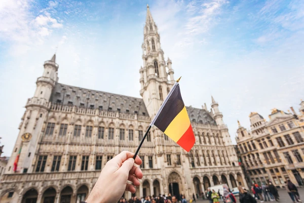 Mano sosteniendo la bandera belga frente a la Grand Place Bruselas, arquitectura gótica icónica y plaza histórica