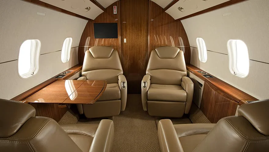 Bombardier Challenger 300 cabin interior forward view showing tan leather seats, wood paneling, oval windows, and entertai...