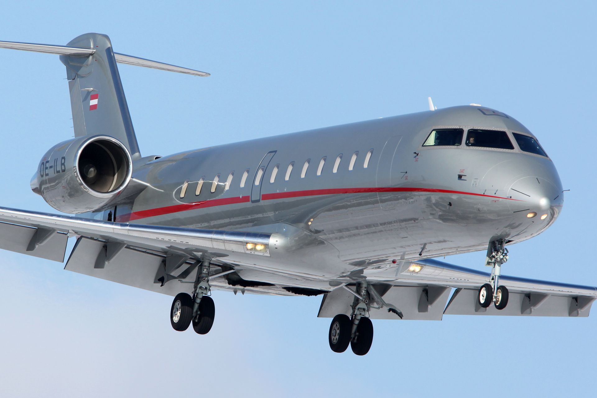 Bombardier Challenger 850 in flight, side view, with landing gear deployed against clear blue sky, showing silver fuselage...
