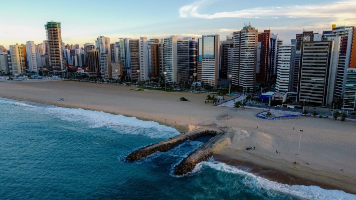 Fortaleza’s beachfront with high-rise buildings along the shore, golden sand, and waves breaking against a stone jetty.