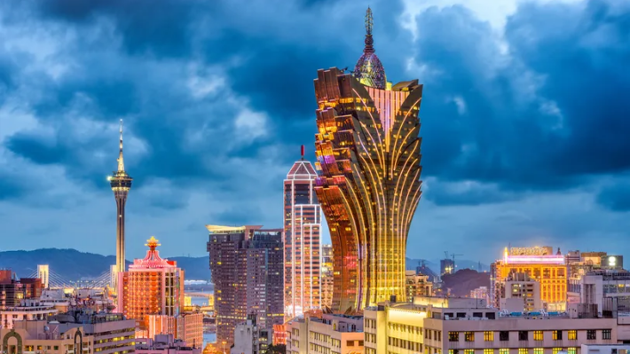 Skyline of Macau at dusk with the illuminated Grand Lisboa tower and surrounding high-rise buildings under dramatic blue clouds.