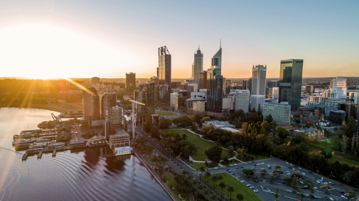 Widok z lotu ptaka na Perth w Australii o zachodzie słońca, ukazujący panoramę miasta, rzekę Swan i otaczające je tereny parkowe, oświetlone ciepłym światłem.