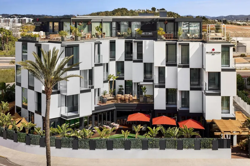 Widok zewnętrzny hotelu Sir Joan na Ibizie, ukazujący nowoczesny, wielopiętrowy budynek z geometrycznymi, białymi fasadami, miejscami do siedzenia na tarasie na dachu i palmami na pierwszym planie.