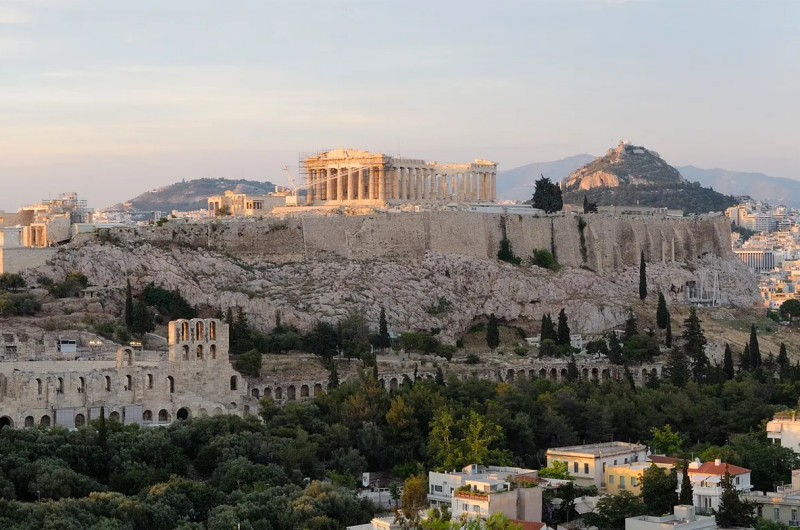 Partenon na Akropolu w Atenach, widziany z daleka o zachodzie słońca, w tle widać miasto i otaczające je wzgórza.