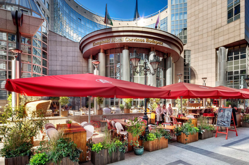 A Kempinski Hotel Corvinus Budapest kültéri terasza piros napernyőkkel, kávézói ülőhelyekkel és a háttérben a szálloda modern üveg- és kőhomlokzatával.
